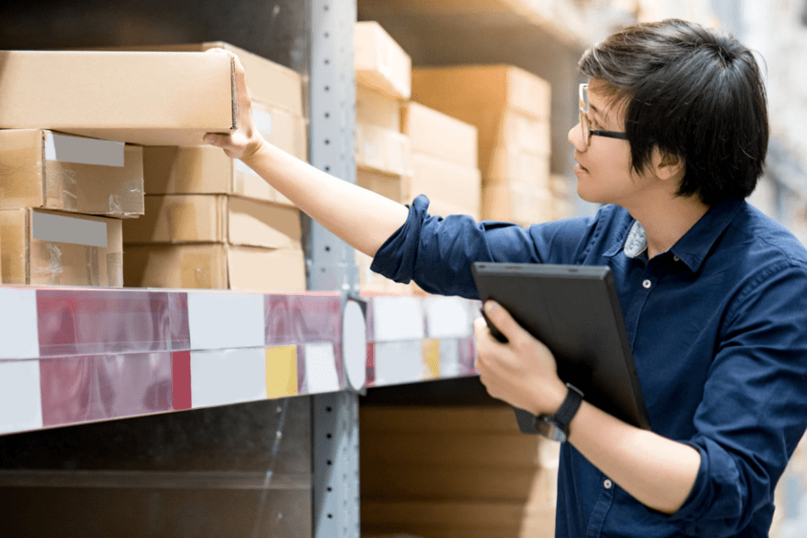 man doing stocktaking of product in cardboard box on shelves in warehouse by using digital tablet