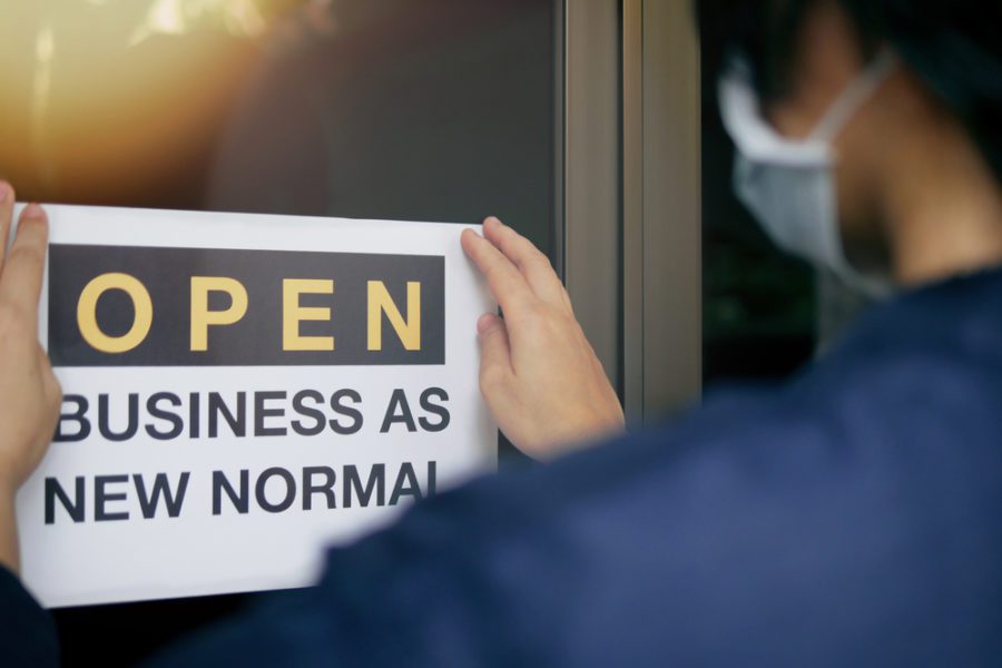 Reopening for business adapt to new normal in the COVID-19 pandemic. Rear view of business owner wearing medical mask placing open sign “OPEN BUSINESS AS NEW NORMAL” on front door.