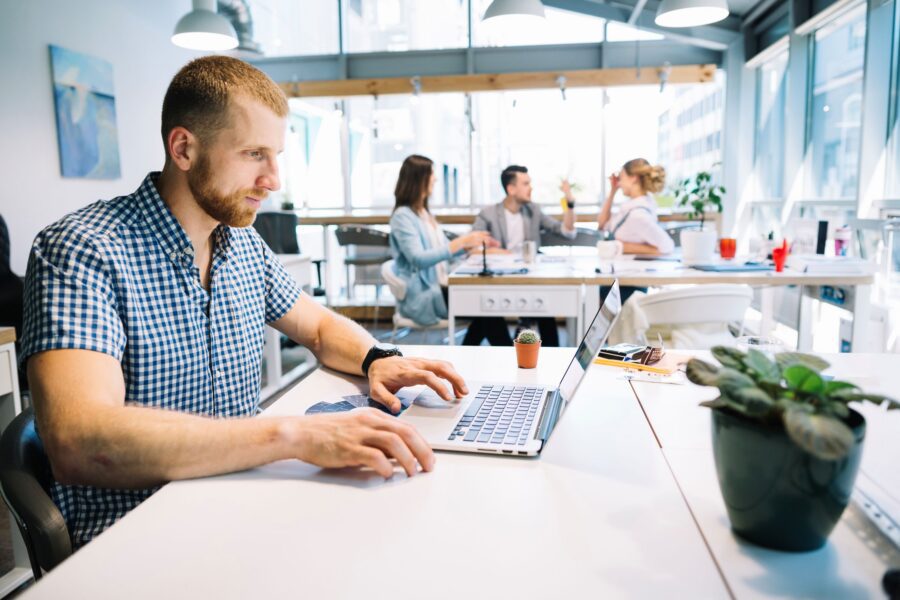 Photograph of a modern office setting with a man in a checkered shirt working on a laptop at a white desk, while three colleagues engage in discussion in the background.