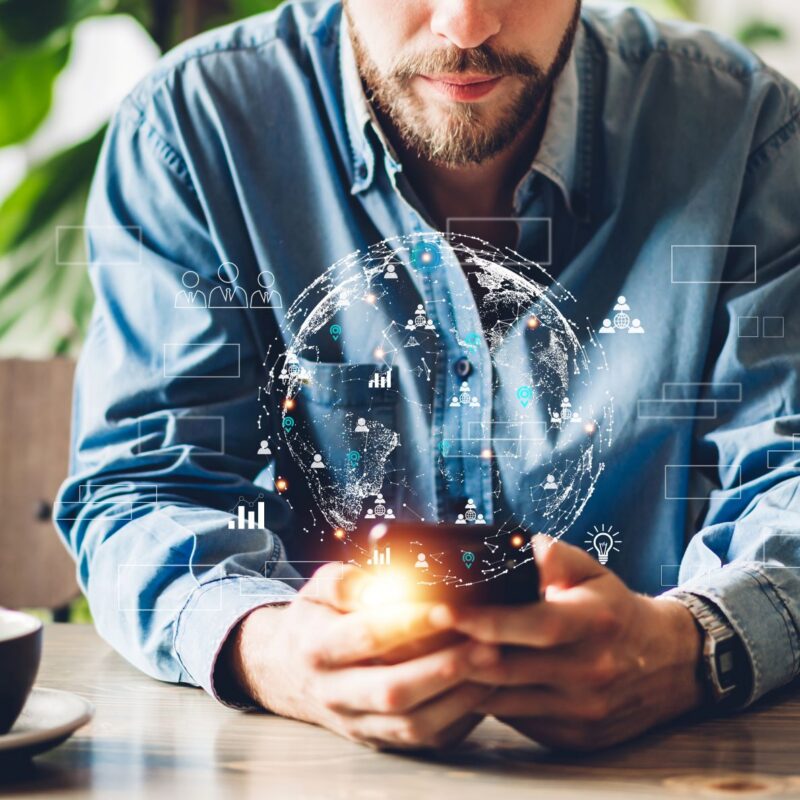 Photograph of a man sitting at a table holding a smartphone with a digital globe and a data visualisation hologram projecting above the device.