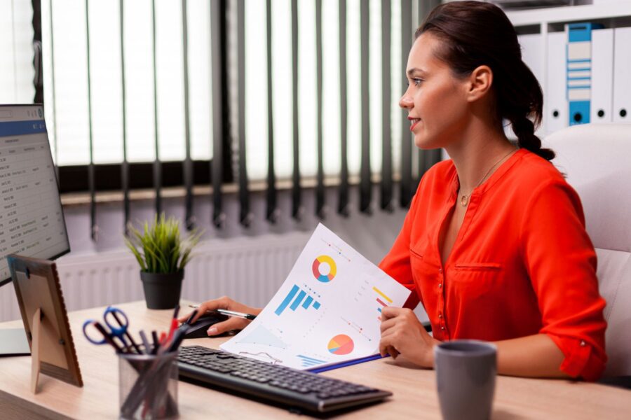 Photograph of a woman working at a desk with a computer and holding a paper displaying colourful charts and graphs.