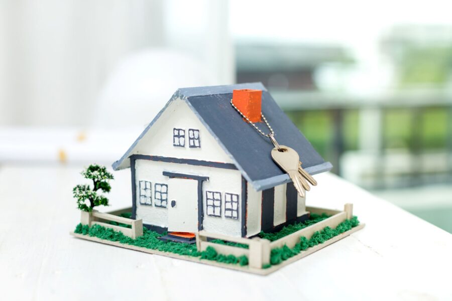 Photograph of a small model house placed on a white surface with a blurred background. A set of silver keys hangs from the chimney, symbolising home ownership or real estate.